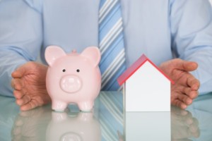 A Piggy Bank Next To A Small House With A Business Man In The Background