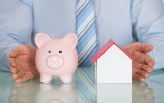 A Piggy Bank Next To A Small House With A Business Man In The Background