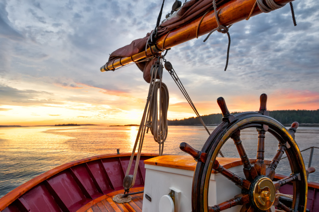 helm - Sunrise sailing on a tall ship schooner. Close up of steering wheel, bow and boom against a dramatic sky at dawn.