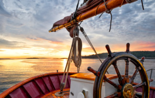 helm - Sunrise sailing on a tall ship schooner. Close up of steering wheel, bow and boom against a dramatic sky at dawn.