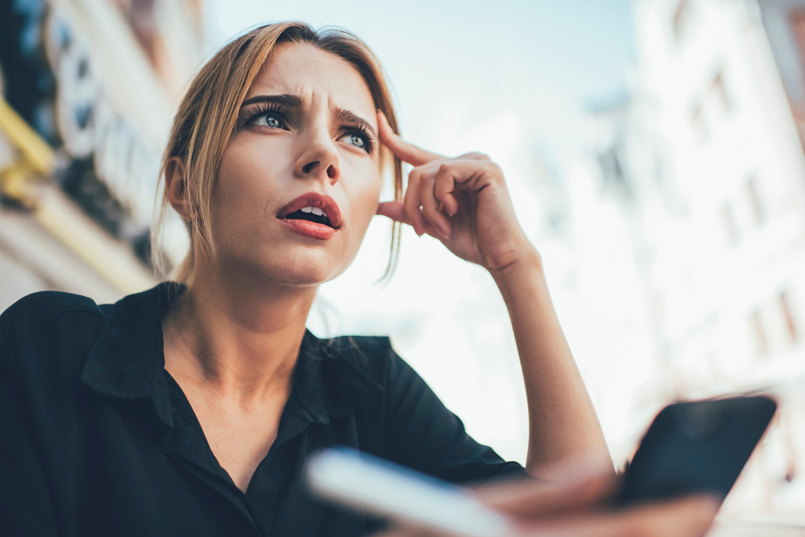 From below of confused blond haired female entrepreneur in black shirt looking away in though while solving problems with partner on mobile phone at city street